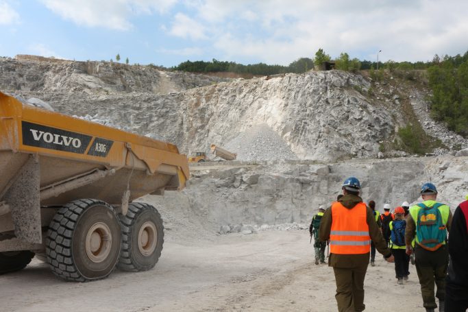 Bagger und Gruppe von Menschen in einem Steinbruch, umgeben von felsigem Terrain.