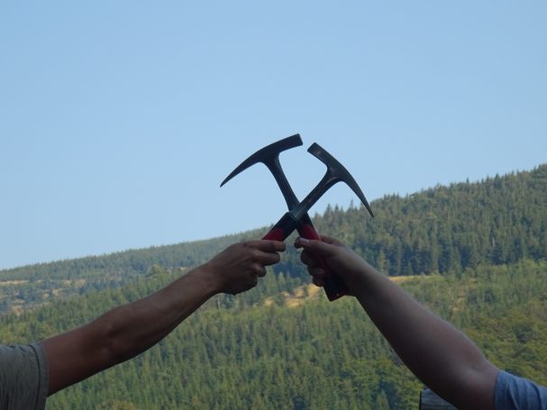 Zwei Personen halten sich Hacken in die Höhe, vor einer bewaldeten Landschaft.